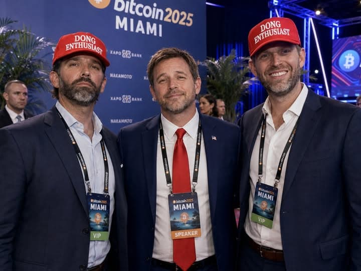Satirical event photo of three men in suits posing at Bitcoin 2025 Miami. The men on either side wear red caps reading “DONG Enthusiast” and VIP badges; the man in the center wears a red tie and a speaker badge.